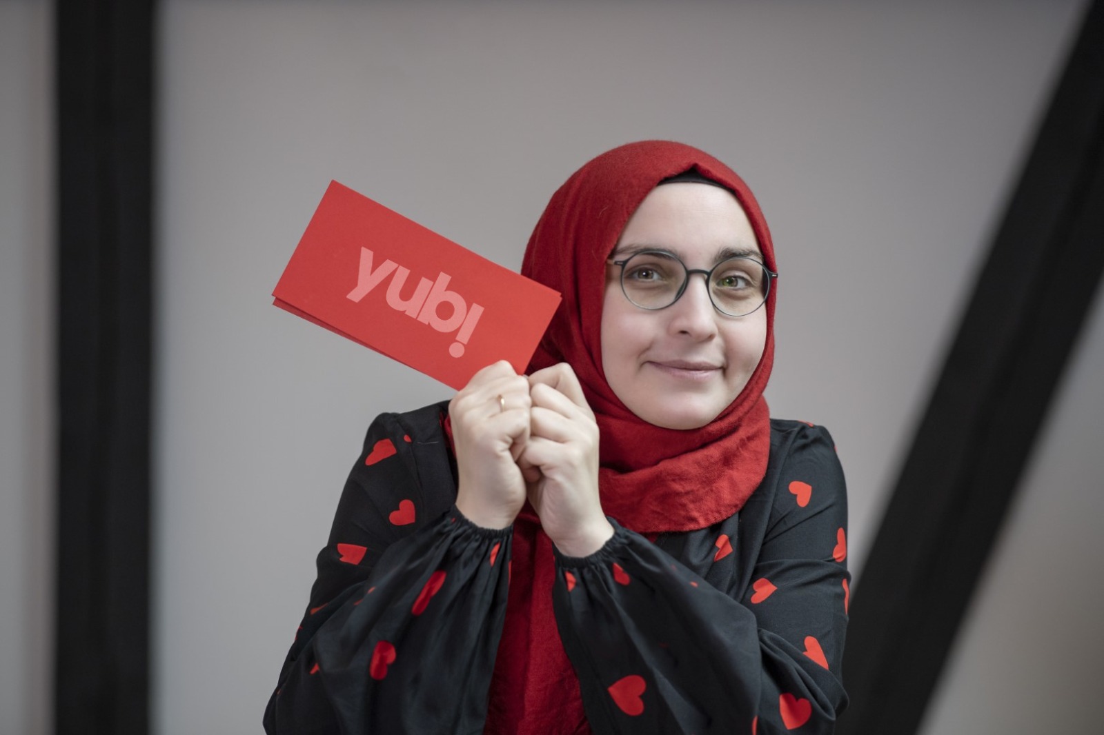 Eine Frau mit rotem Kopftuch und Brille hält lächelnd eine rote Karte mit der Aufschrift "Yubi" in die Kamera. Sie trägt eine schwarze Bluse mit roten Herzen.